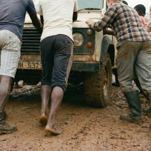 People pushing a truck stuck in the mud on a rural road in Ghana, working together to move it forward.