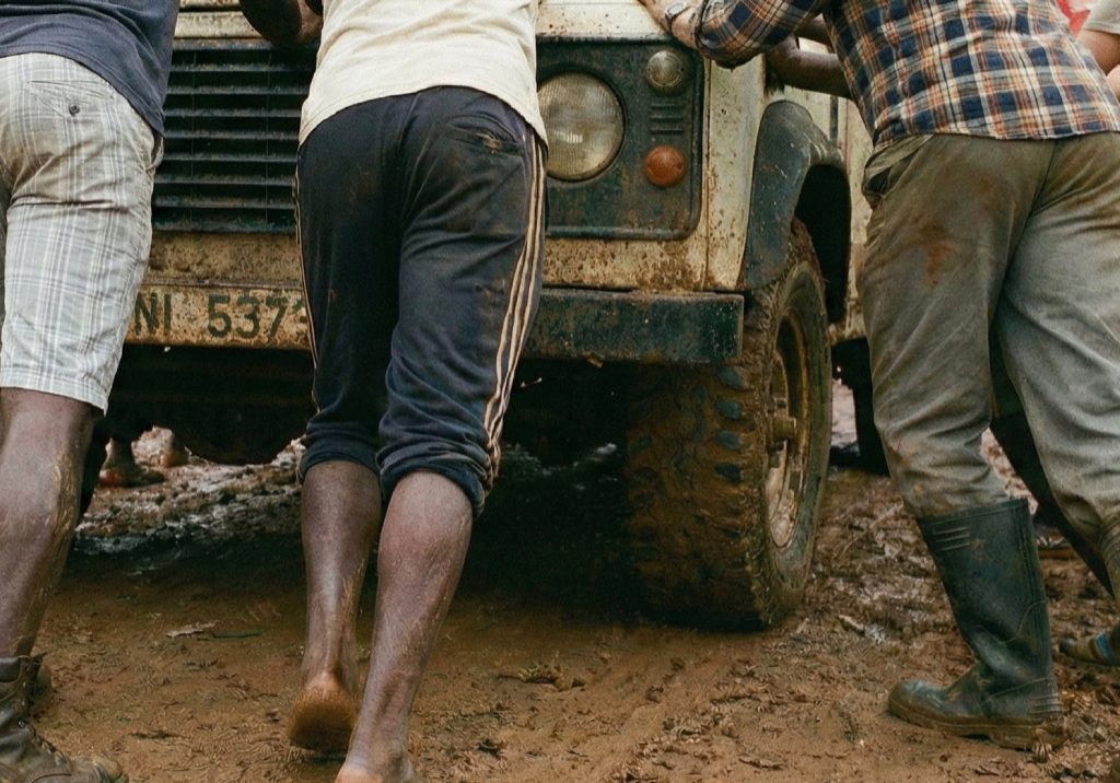 People pushing a truck stuck in the mud on a rural road in Ghana, working together to move it forward.